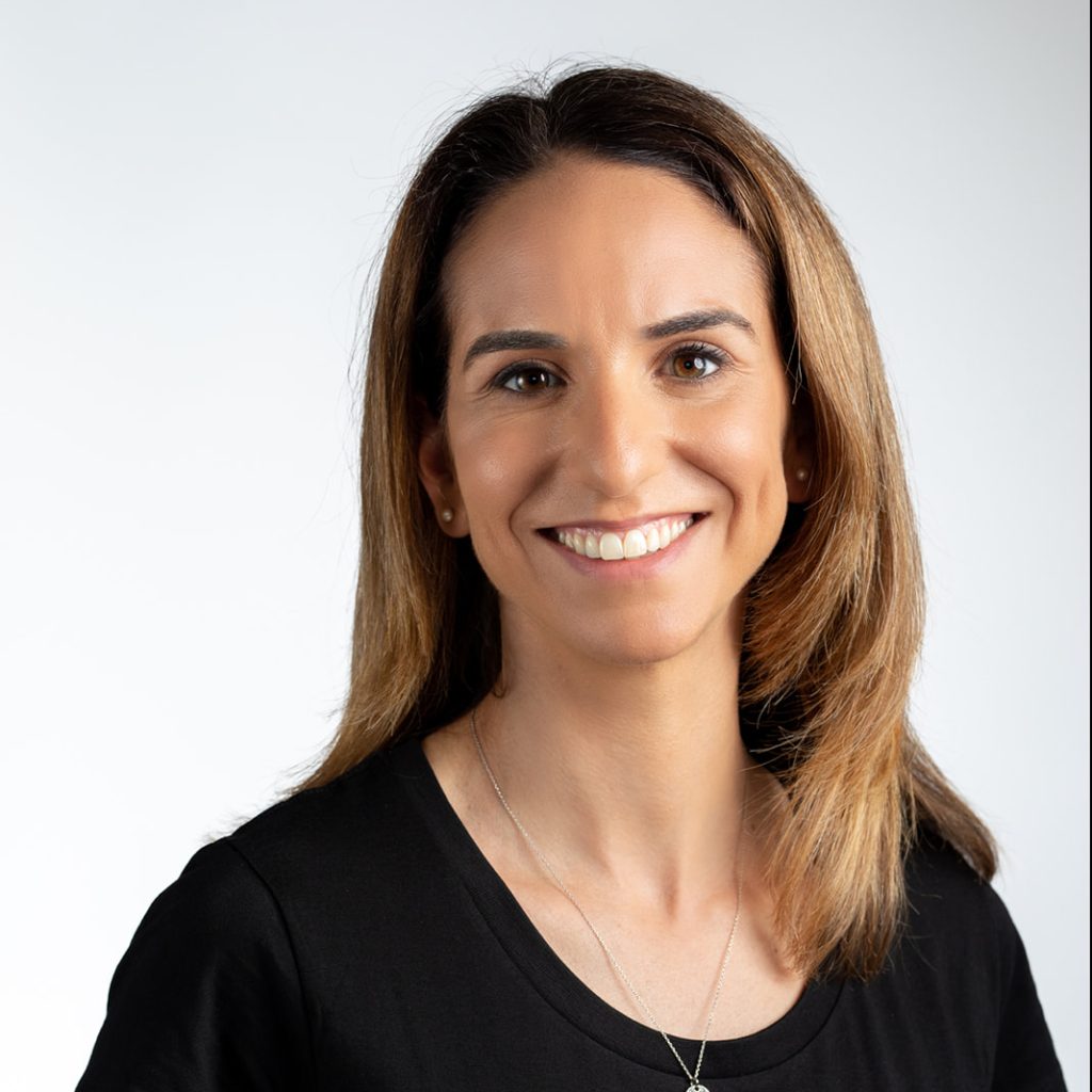 Tamar Schapira is a mid aged, brown haired woman smiling and wearing a black t-shirt with the SenseIT logo on the left hand side.