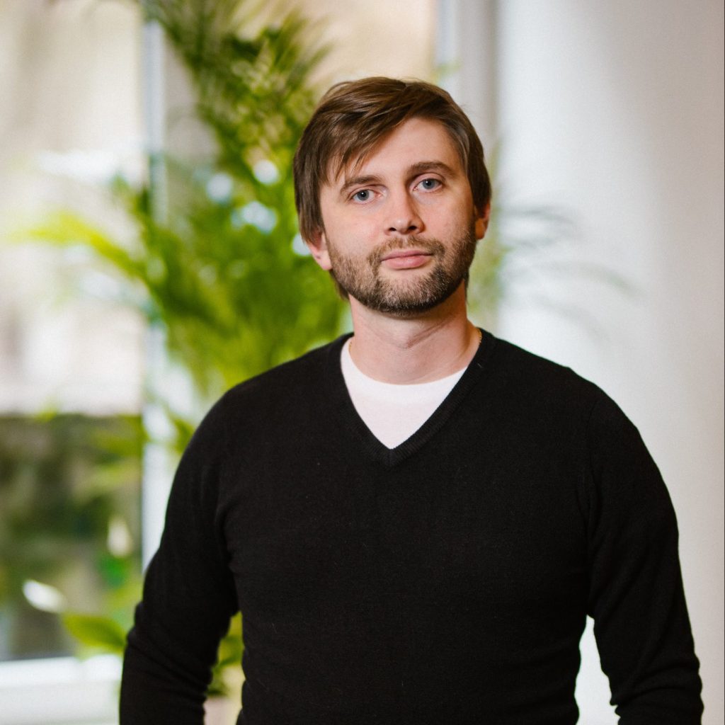 Peteris is a white man with medium brown hair and short beard. He is standing in front of the windowsill on which is a large, green houseplant.