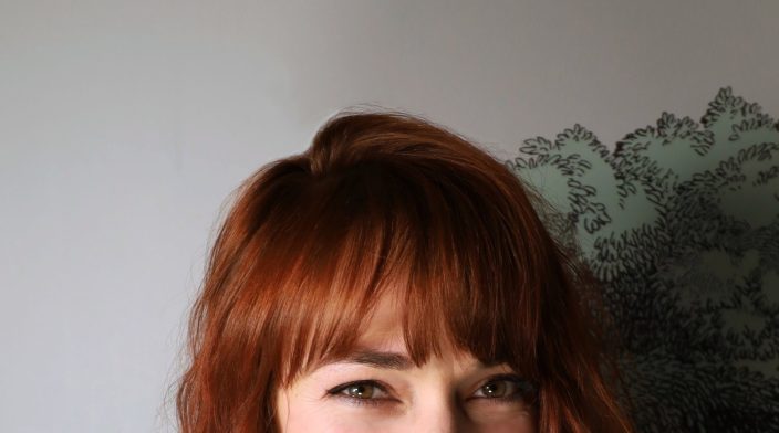Caroline is a white woman in her 30's with wavy auburn hair, bangs, and freckles.