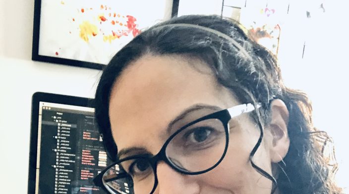 Joni Halabi is a white woman in her 40s with curly dark hair and glasses. She is sitting facing away from a desk with an open laptop whose screen is displaying SCSS code in Visual Studio.