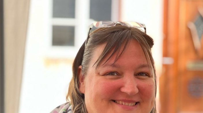Birgit Olzem is a white woman in her 40's with short brown hair and a big smile. Wearing reading glasses like a tiara and a white scarf with different colored dots.