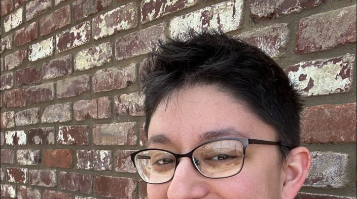 Danielle is standing in front of a brick wall wearing glasses and has short dark hair shaved on the sides. They are wearing a black and red plaid shirt under a heather blue sweater with sunglasses hanging off.