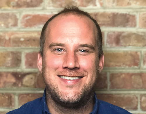 Hans Skillrud is a white man in his late thirties who has a big smile but not a lot of hair on the top of his head (He likes to think he's not 'losing hair', rather he's 'growing face'). Hans has blue eyes and is wearing a blue long sleeve shirt and is sitting indoors in front of a brick background.