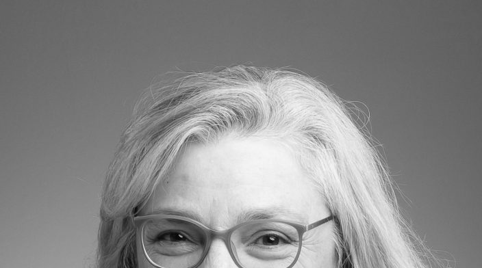 Sandi Gauder is a woman with long silver hair wearing glasses and smiling at the camera.
