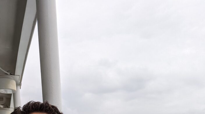 Jason is a white man with short brown hair and a short beard. He is looking over the side of a ferry heading to shore with a smile on his face.