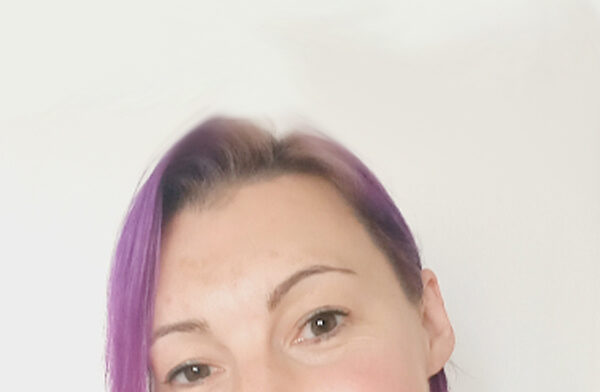 Laura Hutton is a white woman in her 40’s with mid-length purple/pink hair with a friendly smile.