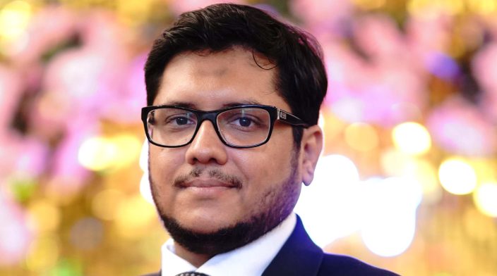 Ali Shan is wearing a dark suit and glasses, and is standing in front of a bright colourful background.