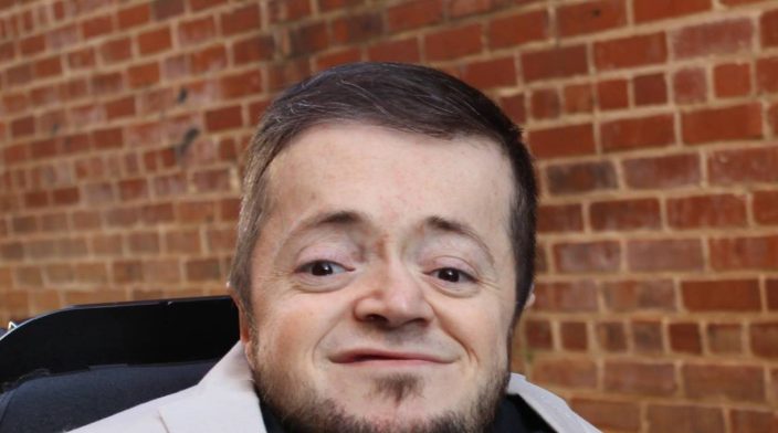 A man with disability wearing a tan blazer and black shirt in-front of a brick wall.