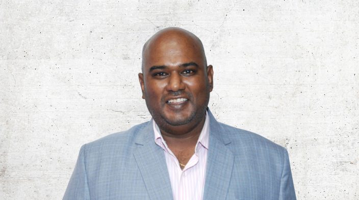 Alexander Gounder is wearing a blue-grey jacket and a pale shirt. He is looking at the camera and smiling,