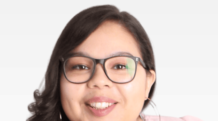 A young woman with long brown hair and glasses sits at a three-quarter angle and rests her chin on her hand.