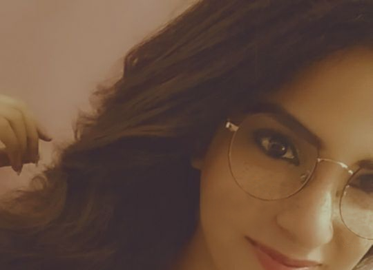 Angled photo of a young woman with long brown hair, smiling, and wearing glasses.