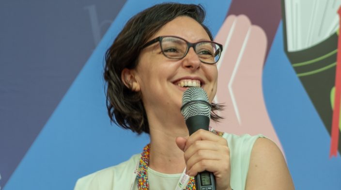Alice Orrù is a white woman in her 40s; she has brown bob hair, large tinted-framed glasses and a joyful smile. She holds a microphone and has on her lap the book she wrote on inclusive languages in Italian.