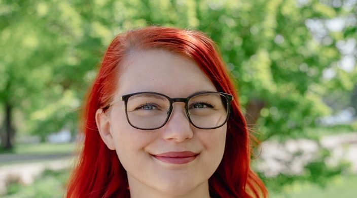 Tina Reis is a white non-binary person in their early 30s with medium-long red hair and glasses. In the photo, they are wearing a blue blouse and there is a park with green trees in the background.