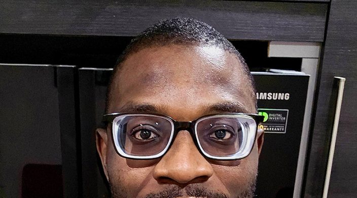 David is a Black African man in his early 30's wearing a black pendant necklace on an army green t-shirt and a pair of thick medicated glasses.