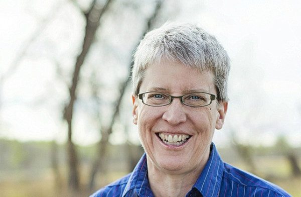 Lorraine Watson is a white woman with short grey hair, glasses, and big smile standing in front of trees in a park