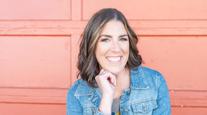 Sarah Kornblum is a woman with shoulder length brown hair. She is wearing a jean jacket and is in front of an orange background.