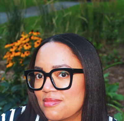 A black woman wearing thick framed black glasses and straight brown hair, she is wearing a starkly contrasted white and navy striped dress. She is sitting in a garden with black-eyed Susans in the background.