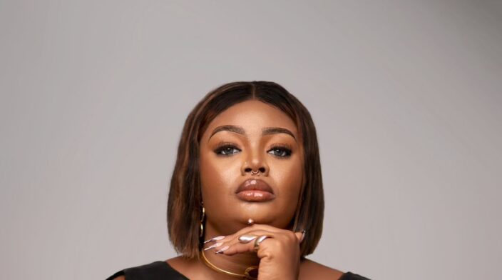 Victoria Ottah is an African lady from Nigeria. She has type C hair type but is currently in a short straight brown wig. She is sat in a professional pose, in a black dress and arms folded to her jaw.