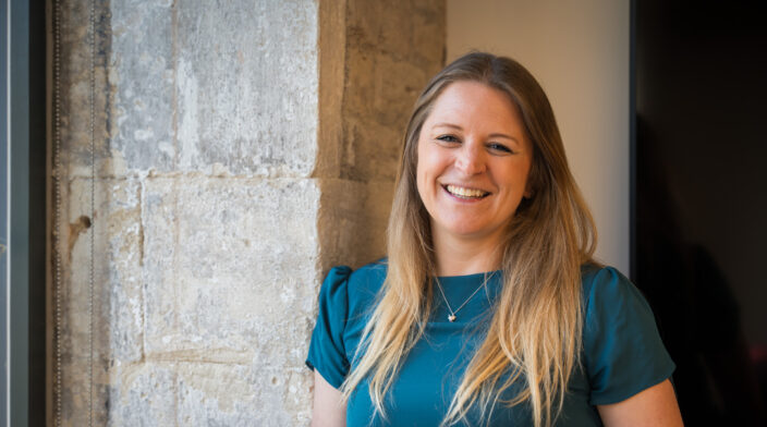 Lucy Collins, a white woman in her mid-30s with long blonde hair, leans against a pale stone wall at Web Usability HQ. She wears a teal t shirt and has a big smile on her face.