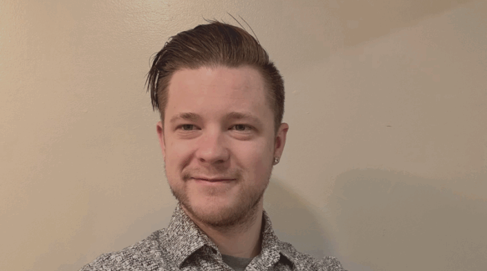 Shown from the chest up, James standing with his arms crossed and slightly angled away from the camera. James is wearing a button up shirt that is black and white. His brown hair is swooped over away form his face and he is clean shaven. James smiles while looking back at the camera.