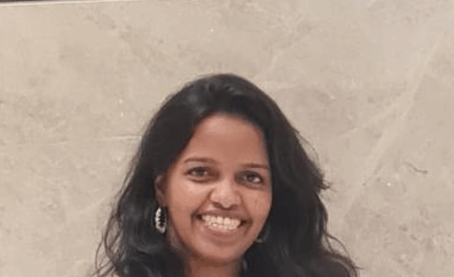 Janaki, smiling with long hair, wearing a light pink top, standing in front of a light background.