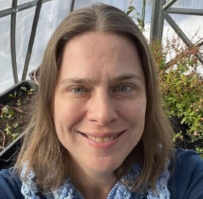 Elaine, a white female with shoulder-length dirty blonde hair, stands smiling in her greenhouse in front of a leafy rosebush and a sparsely planted box of spinach. She is wearing a blue top and crocheted sky blue neck warmer.