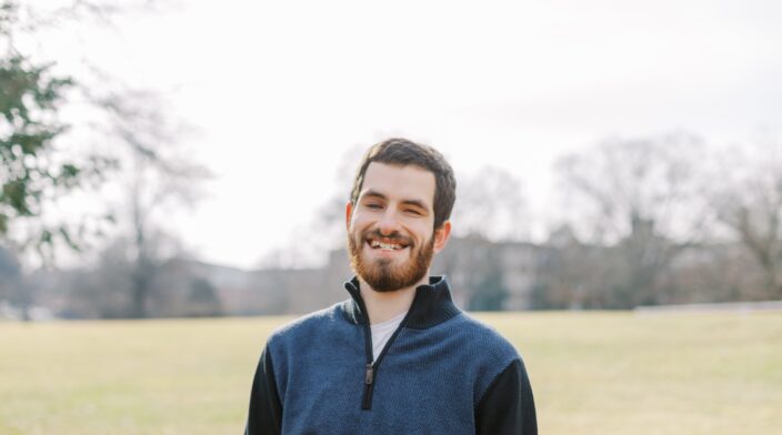 Kevin, a white male with brown hair and a short beard, stands smiling in a brightly lit field. He is wearing a blue and black quarter-zip sweater and looks relaxed.