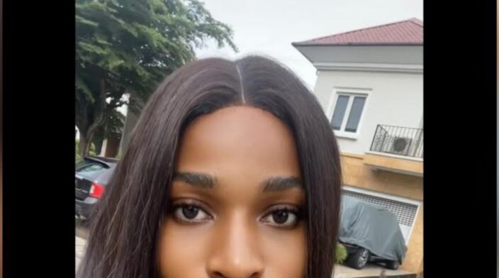 Yigakpoa Ikpae is an African woman with brown skin. She took a selfie while wearing a burnt orange dress, short straight weave and a slightly visible gold chain. Behind her is a building, green tree and two cars.