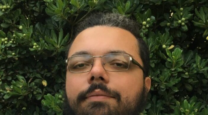 Mycke Ramos is a latino man in his 30s with curly black hair tied back. In their background you can see leaves from a tree.