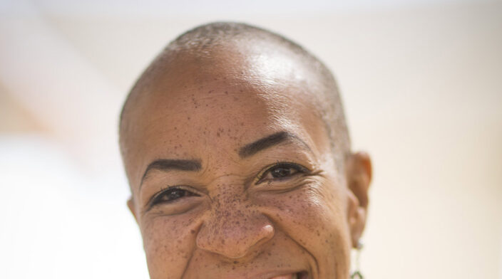 Bronze skinned woman with freckles and closely shaved head, smiling on a sunny day.