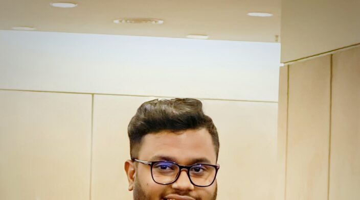 Nasim Miah is a young South Asian man with short dark hair, a trimmed beard, and glasses. He is smiling warmly while standing in front of a microphone at WordCamp Malaysia 2023 Contributor Day. He is wearing a dark patterned shirt with the word ‘Guess’ on it and a blue lanyard.