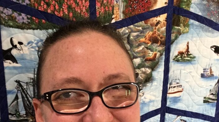 Headshot of Amy Carney, a smiling middle-aged freckled white woman wearing dark-framed plastic glasses with thick lenses. Behind her is a sashed quilt with scenes from Alaska.
