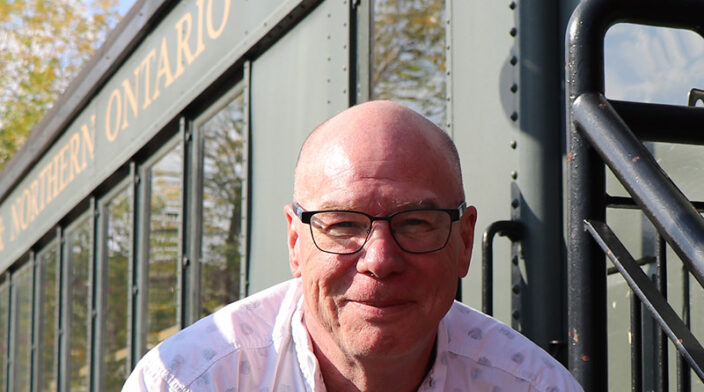 An older male with a white skin tone wearing a white button-up shirt sitting in front a old style green train car on a sunny day.