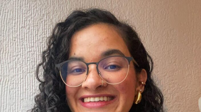Alexandra Blanco is a hispanic woman in her mid-20s with long curly black hair. She wears glasses and a white blouse with golden jewelry on her ears and nose.
