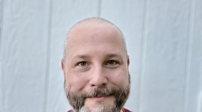 Chris Hinds is a white man in his late 30s with a shaved head and full, neatly trimmed beard. His hair is a mix of brown and gray and he is wearing a red t-shirt. He is smiling slightly and looking directly at the camera.