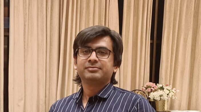 Dilip is an Indian man in his 30s with short black hair and glasses. He is smiling and wearing a collared shirt, standing in front of a plain background.