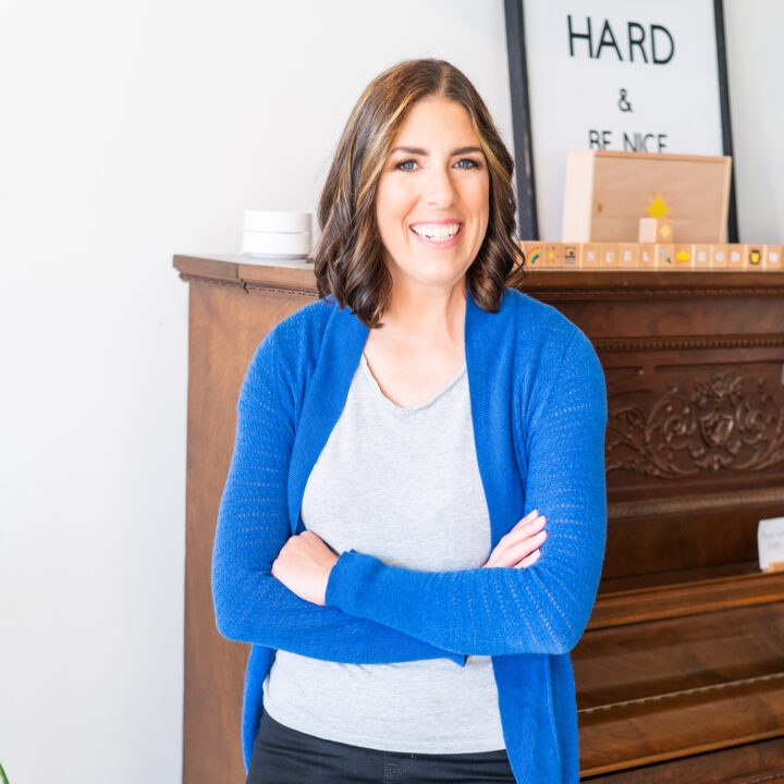 Sarah Kornblum smiling and wearing a vibrant blue cardigan standing in front of a piano