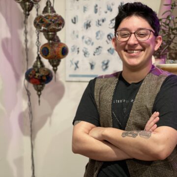 Danielle has short dark hair and is smiling at the camera with arms crossed, wearing glasses, a vest, and short sleeved shirt, and a robot tattoo is partially visible on their forearm.