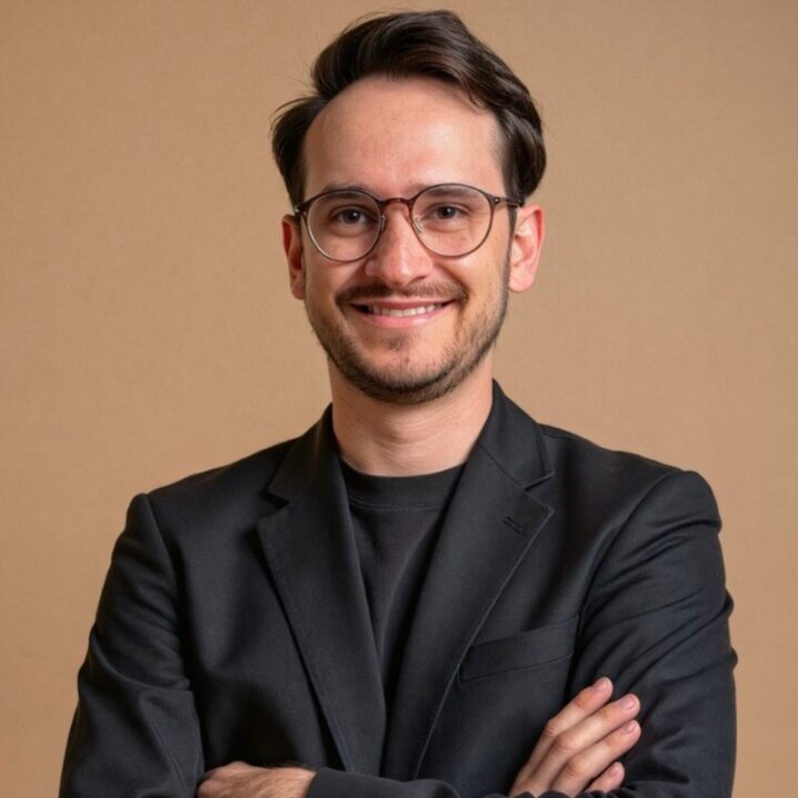 Adam Unger is a man with short dark brown hair swept back, a short beard, and round tortoiseshell glasses. He is smiling with his arms crossed, wearing a black blazer over a black t-shirt.
