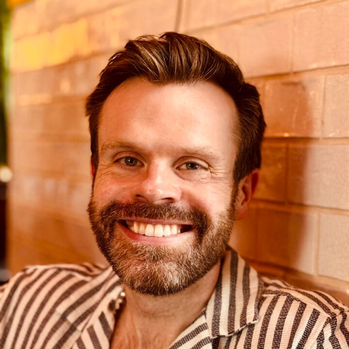 Carl is a white man in his 30s with neatly styled brown hair, a full trimmed beard, and a broad, friendly smile. He is wearing a striped collared shirt and is photographed in warm indoor lighting against a brick wall background.