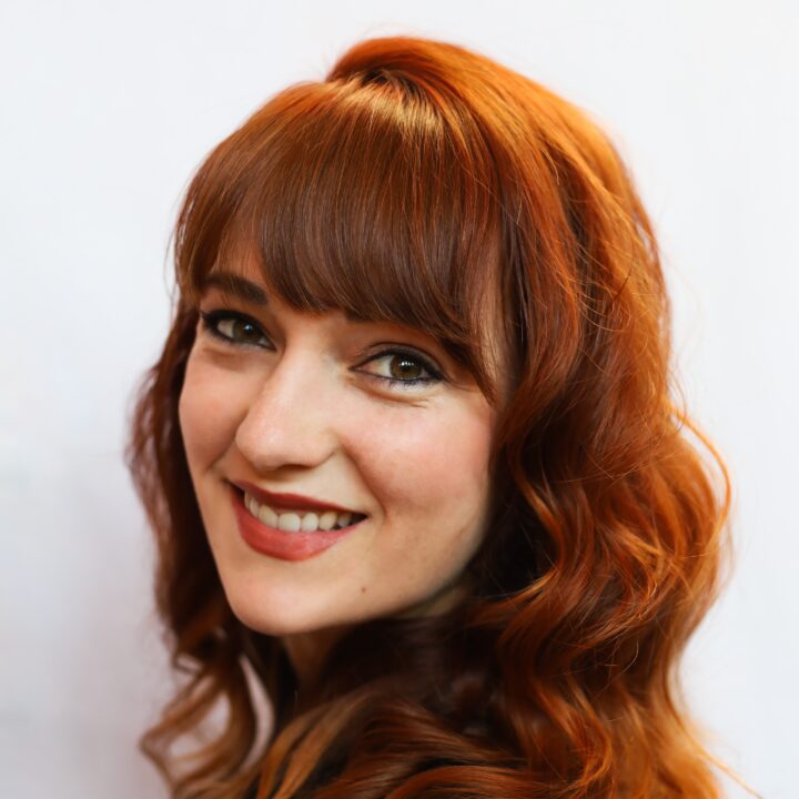 Headshot of Caroline Desrosiers, a white woman in her 30s with wavy auburn hair, bangs, and freckles turning to look at us with a warm smile.