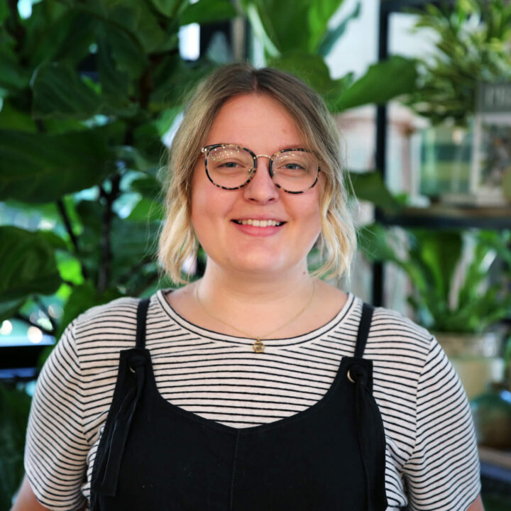 A white woman in her 30s stands smiling in a bright plant shop. She has short, light brown hair, round glasses, and wears black overalls over a black-and-white striped shirt.