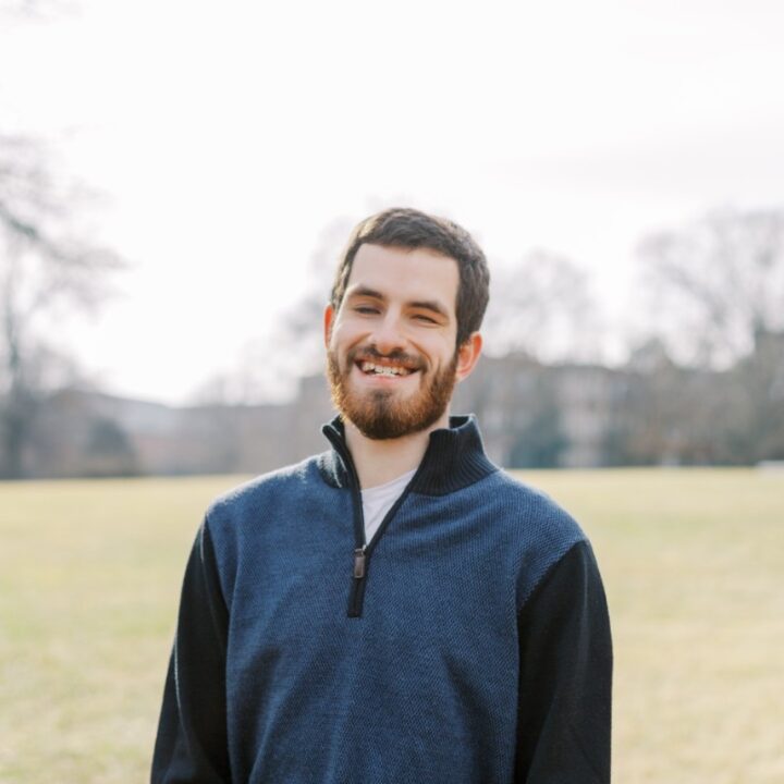Kevin, a white male with brown hair and a short beard, stands smiling in a brightly lit field. He is wearing a blue and black quarter-zip sweater and looks relaxed.