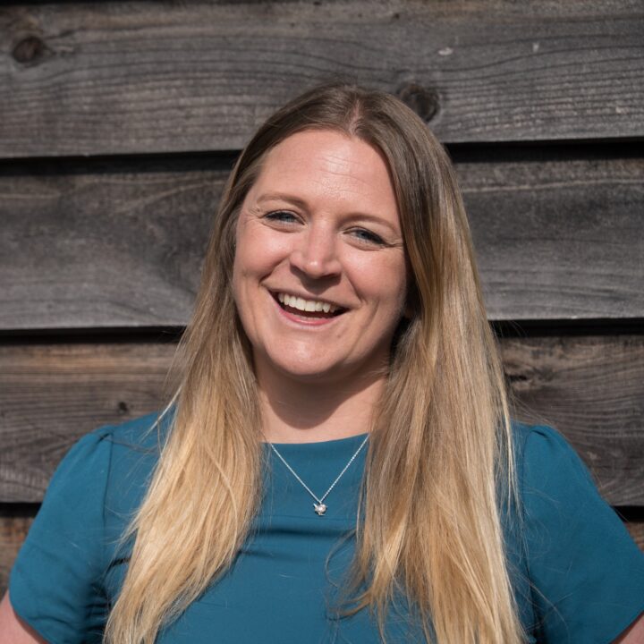 Lucy is a white woman in her mid 30s with long blonde hair parted down the centre. She wears a teal t shirt and leans against a wood clad building with the sun shining on her smiley face.