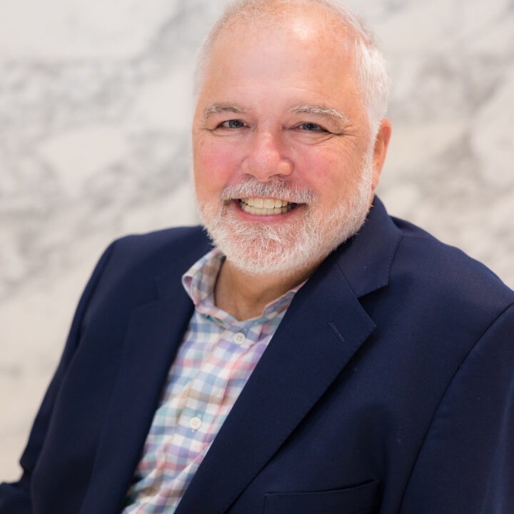 Professional headshot of John Mark Wallach, MBA, smiling while seated, wearing a navy blazer over a pastel plaid shirt, against a softly blurred light marble background.