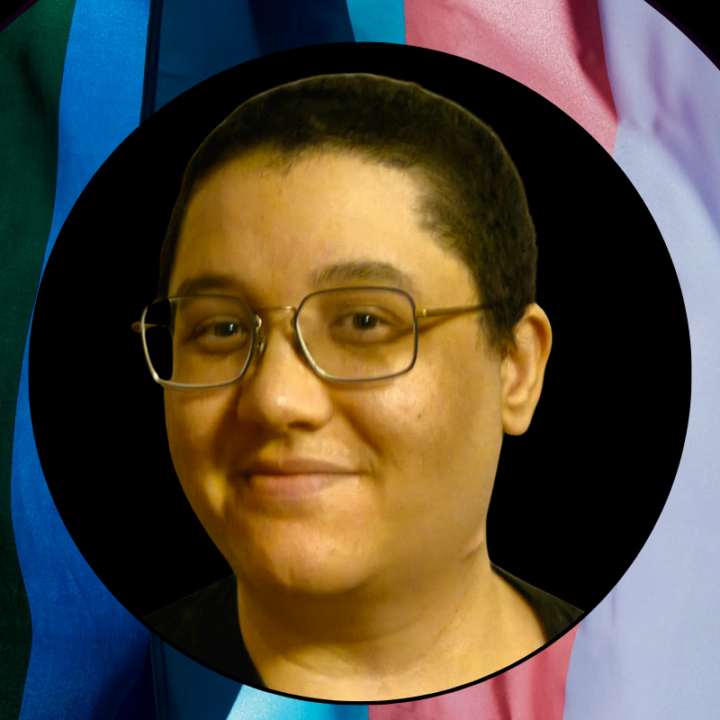 Tas Kronby is a person of color almost bald with black hair. They are wearing a large pair of square glasses. In the background are the pride and trans flag colors.
