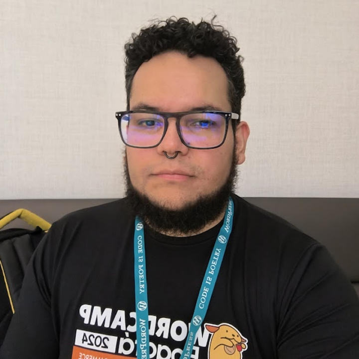 A person with light-to-medium skin, short dark curly hair, black rectangular glasses, and a neatly trimmed full beard with a connected mustache. They are wearing a black WordCamp Madrid 2024 T-shirt featuring a cartoon cat, a blue "Code is Poetry" WordPress lanyard, and a black septum ring. They are seated indoors against a neutral-colored wall with a calm, neutral expression.