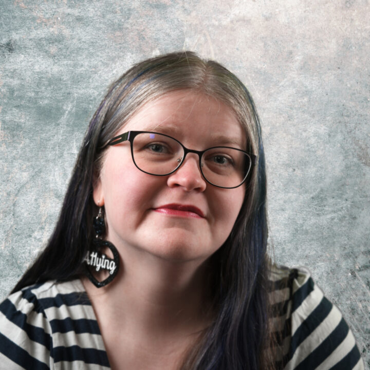 Sanna is a white woman with long, dark ashy blonde hair, which is parted in the middle and highlighted with blue and purple on both sides. She wears black-rimmed glasses and red lipstick, and has a gentle smile. She is wearing a blue-and-white striped top and large, black, heart-shaped earrings that displays the word 'A11ying' in script. She is posed against a textured, mottled grey-blue background.