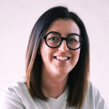 Valentina Mariotti is a white woman in her 40s with straight, shoulder-length dark brown hair and round black glasses. She has a warm, big smile and is photographed against a plain light pink background, wearing a white t-shirt.