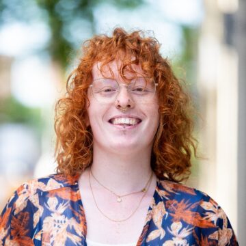 Autumn Rockwell is a white, non-binary person with short, red curly hair and glasses. They are in their early 20's, and they have several facial piercings.
